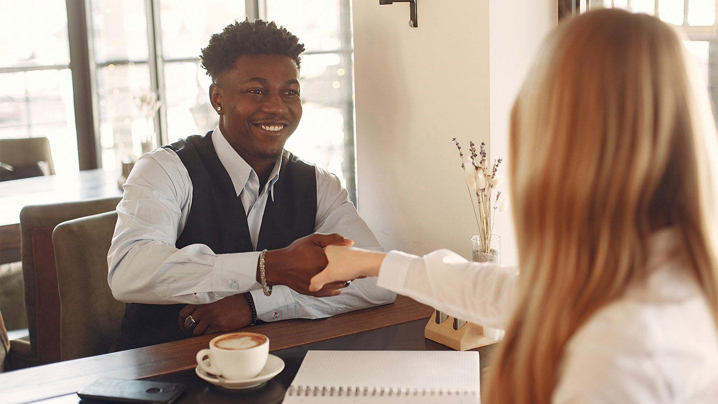 an interviewee sat at a table, smiling whilst shaking hands with an interviewer