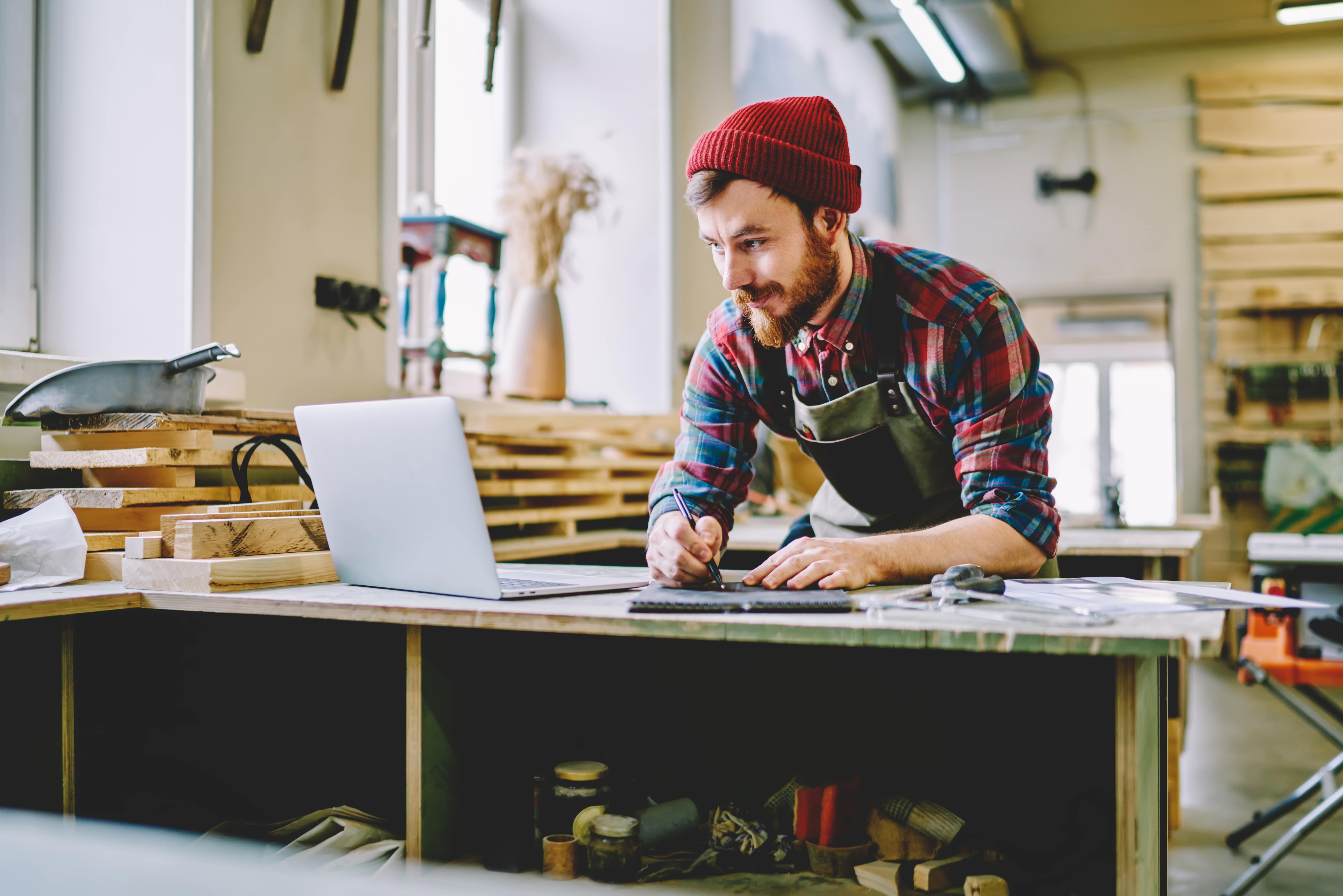 Bearded man in a red beanie and plaid shirt using a laptop and taking notes in a woodworking workshop