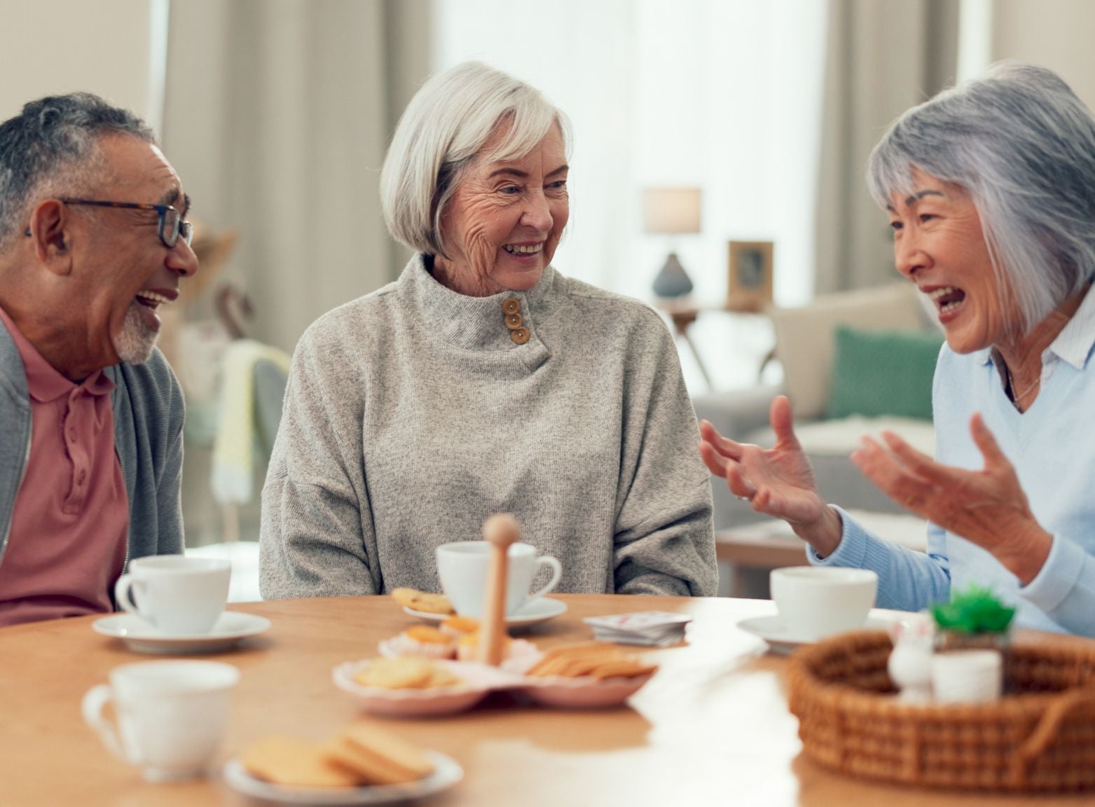 Three older adults engage in conversation over tea and cookies at a cozy table in a bright, modern living room
