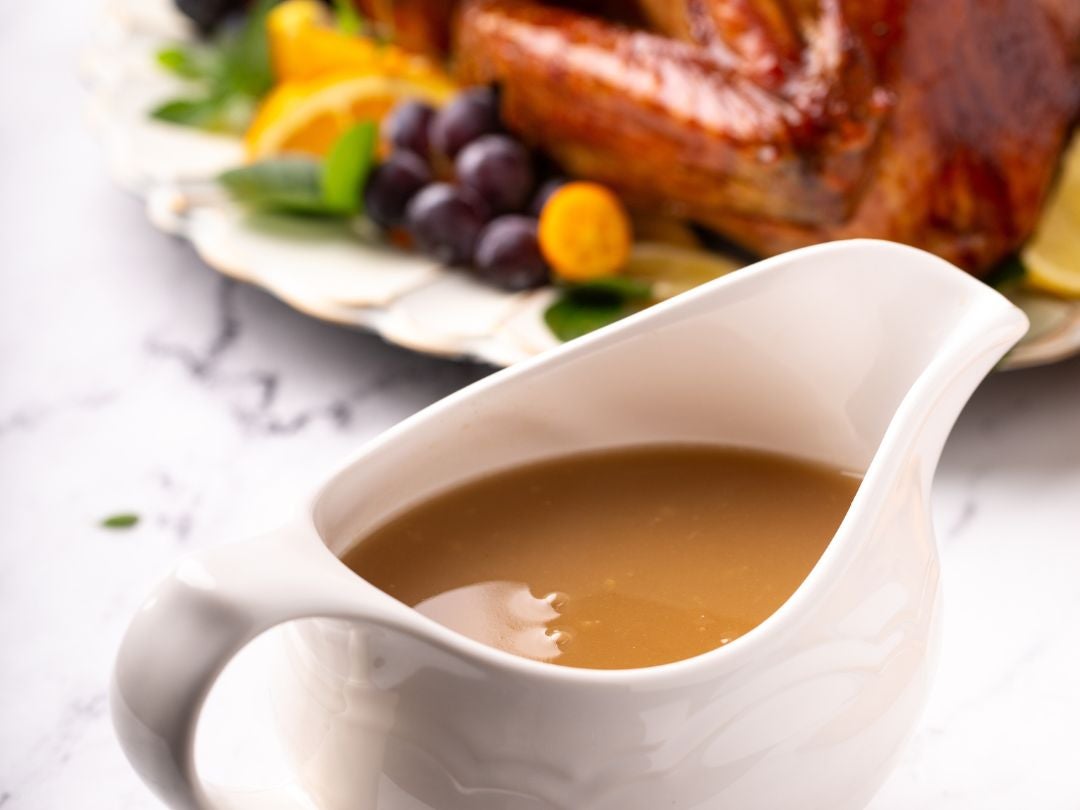A creamy gravy in a white pitcher, with a festive platter of roasted turkey and colorful fruits in the background.