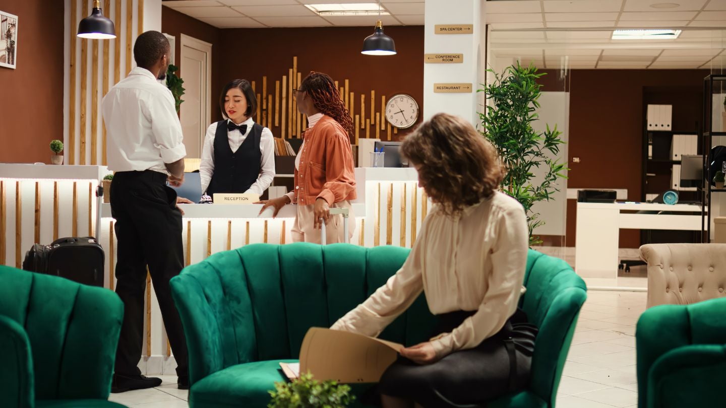 A modern hotel reception area with two guests speaking to a receptionist, while another guest sits on a green couch