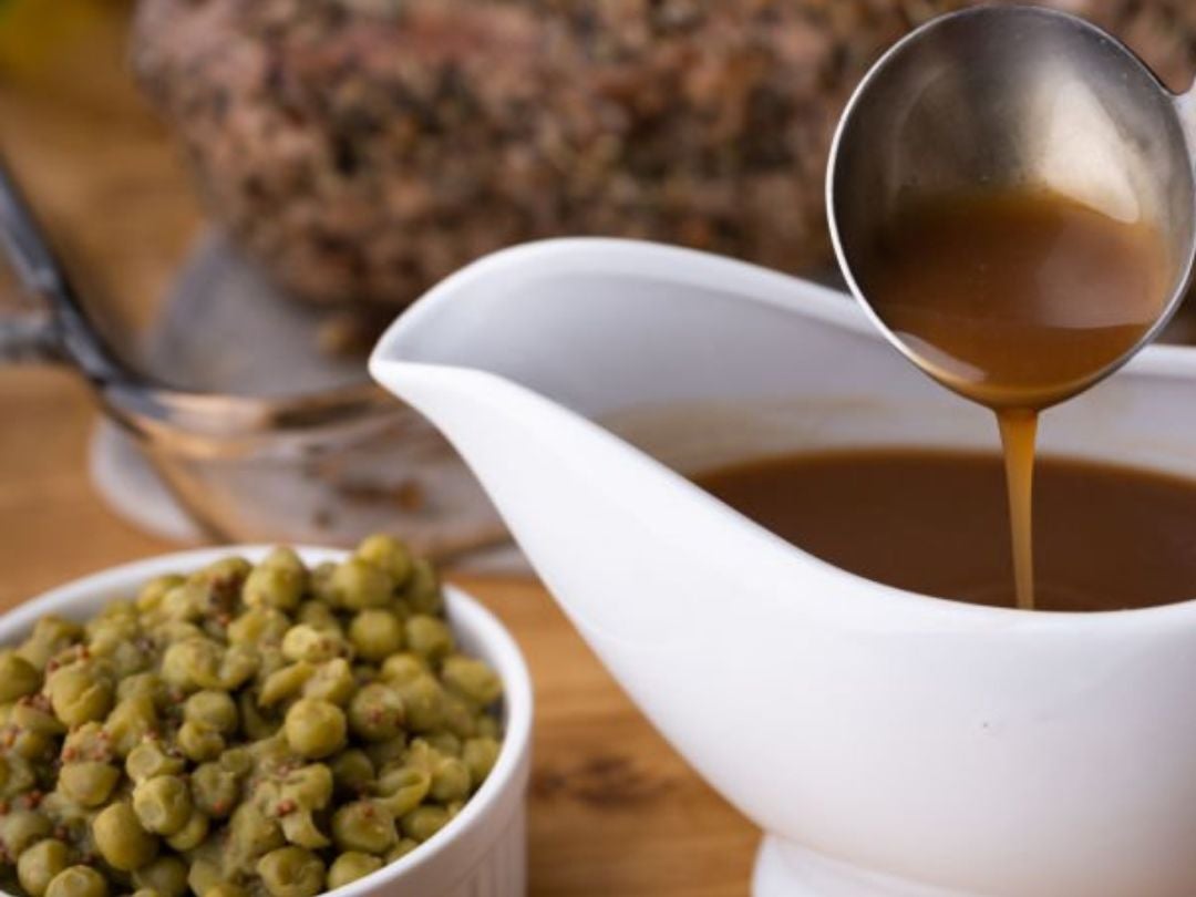 A gravy boat filled with brown gravy, being served, next to a bowl of green peas on a wooden table.