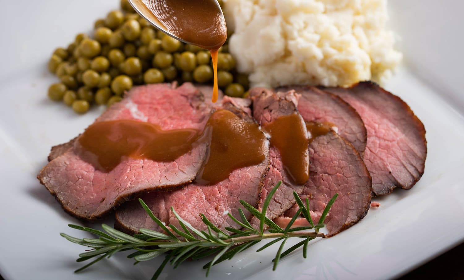 Gravy poured on roast beef with potatoes and peas.