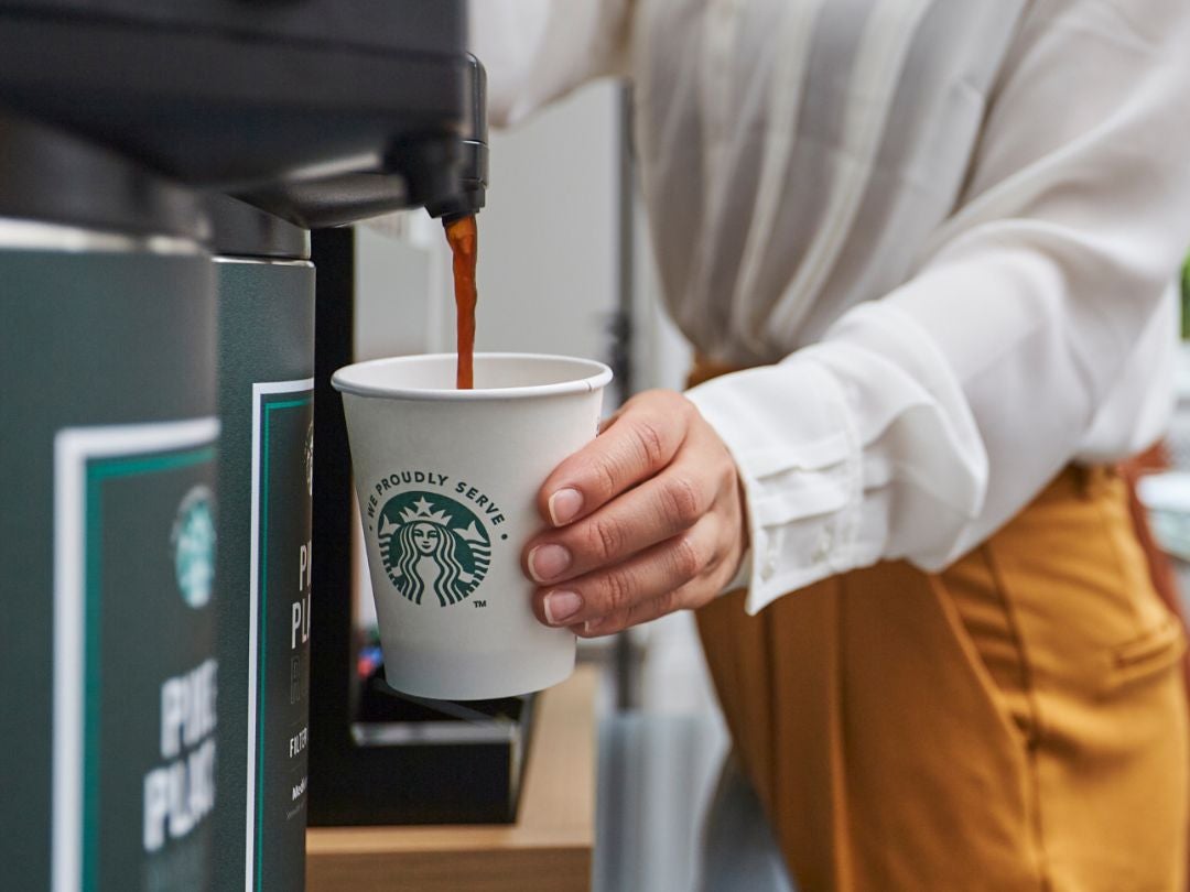A person in a white shirt and brown pants holding a Starbucks cup as coffee is dispensed from a machine.