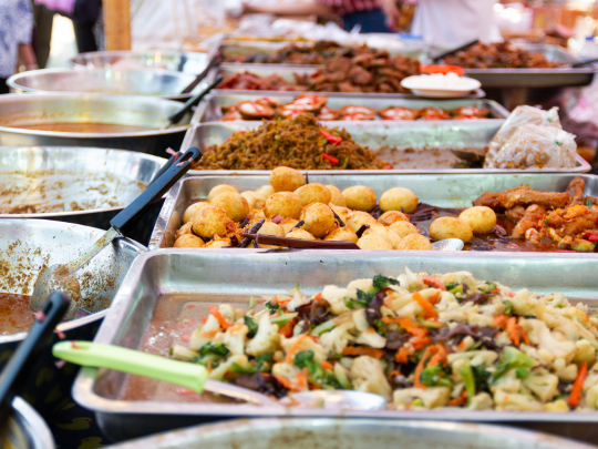 Variety of cooked dishes including vegetables, fried snacks, and rice served in metal trays at a market food stall.
