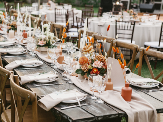 Long wooden table set for event with beige napkins, glassware, floral centerpieces, dried orange accents, and candles