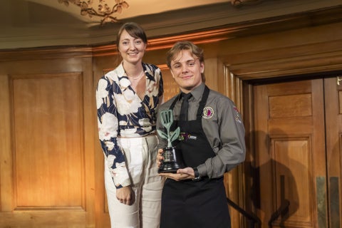 Two people stand indoors posing with a leafy trophy; one wears a floral blouse and white pants, the other a dark apron