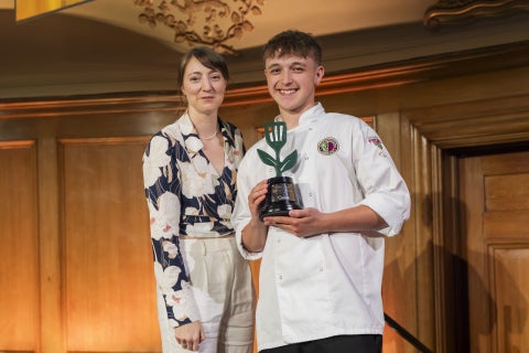 Two people stand on stage, one holding a green trophy shaped like a fork with a leaf design.