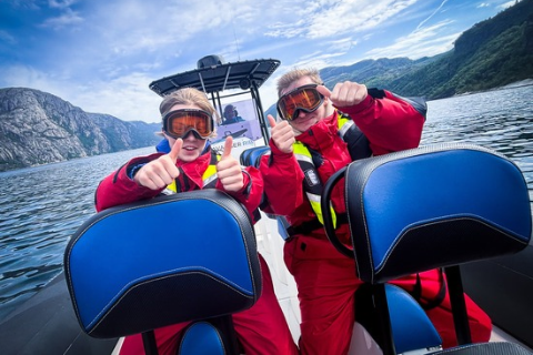 Two people in red dry suits on a speedboat, giving thumbs up on a fjord under blue sky.