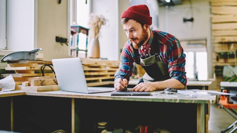 Bearded man in a red beanie and plaid shirt using a laptop and taking notes in a woodworking workshop