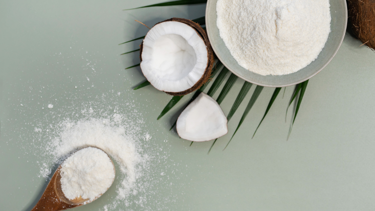 A bowl of coconut powder beside halved coconuts and scattered powder on a light green surface, accented with palm leaves.