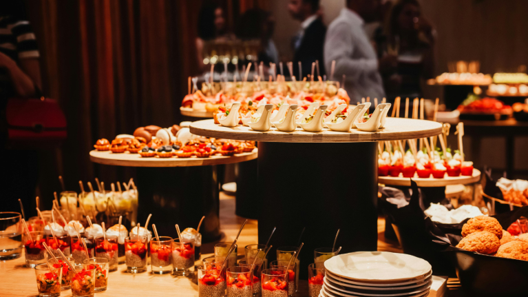 Buffet table with assorted appetizers and desserts in small glasses and plates, set up at an indoor event with blurred guests