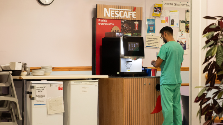 Person in green scrubs using a Nescafe coffee machine in a break room with a clock on the wall showing 3:53.