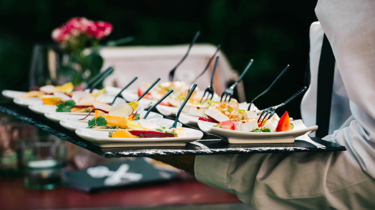 Waiter carrying tray with multiple plates of gourmet appetizers, each with a small fork, against a blurred background.