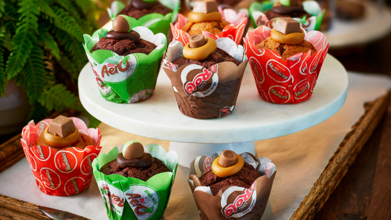 Assorted chocolate and caramel muffins topped with candies, displayed on a white marble stand and wooden tray.