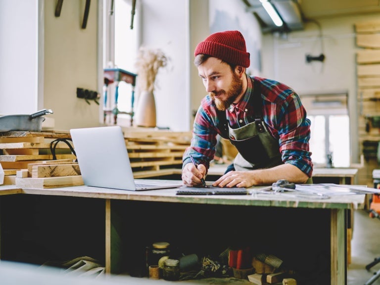 Bearded man in a red beanie and plaid shirt using a laptop and taking notes in a woodworking workshop