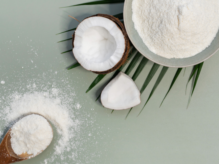 A bowl of coconut powder beside halved coconuts and scattered powder on a light green surface, accented with palm leaves.