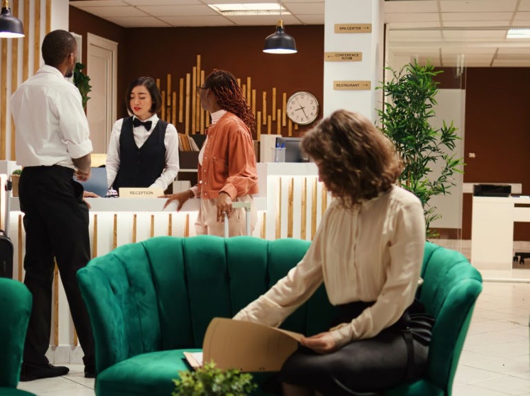 A modern hotel reception area with two guests speaking to a receptionist, while another guest sits on a green couch