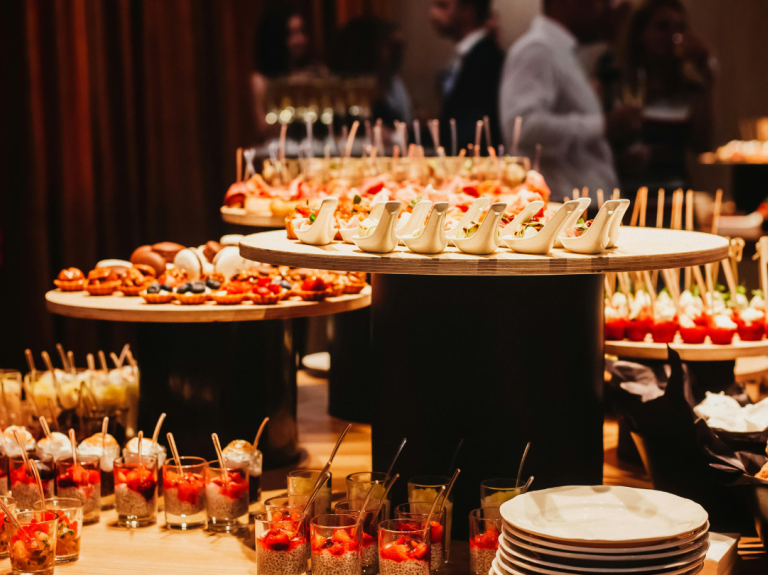 Buffet table with assorted appetizers and desserts in small glasses and plates, set up at an indoor event with blurred guests