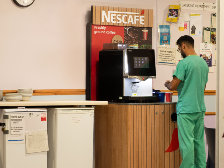 Person in green scrubs using a Nescafe coffee machine in a break room with a clock on the wall showing 3:53.