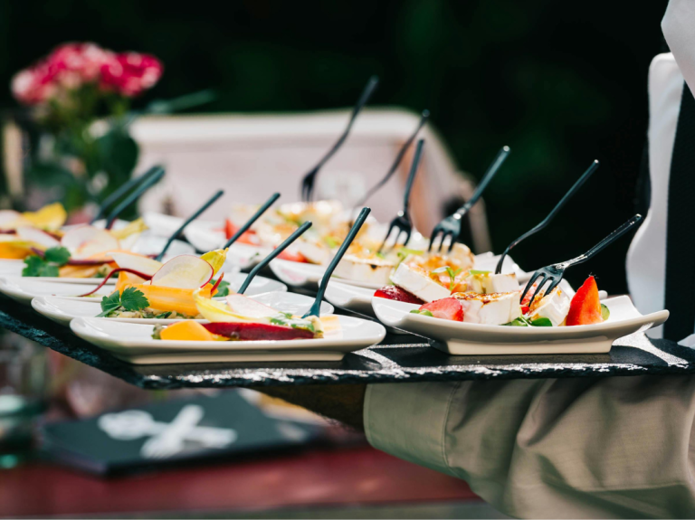 Waiter carrying tray with multiple plates of gourmet appetizers, each with a small fork, against a blurred background.