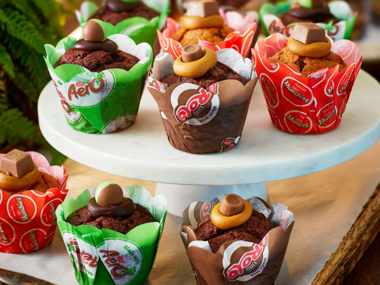 Assorted chocolate and caramel muffins topped with candies, displayed on a white marble stand and wooden tray.
