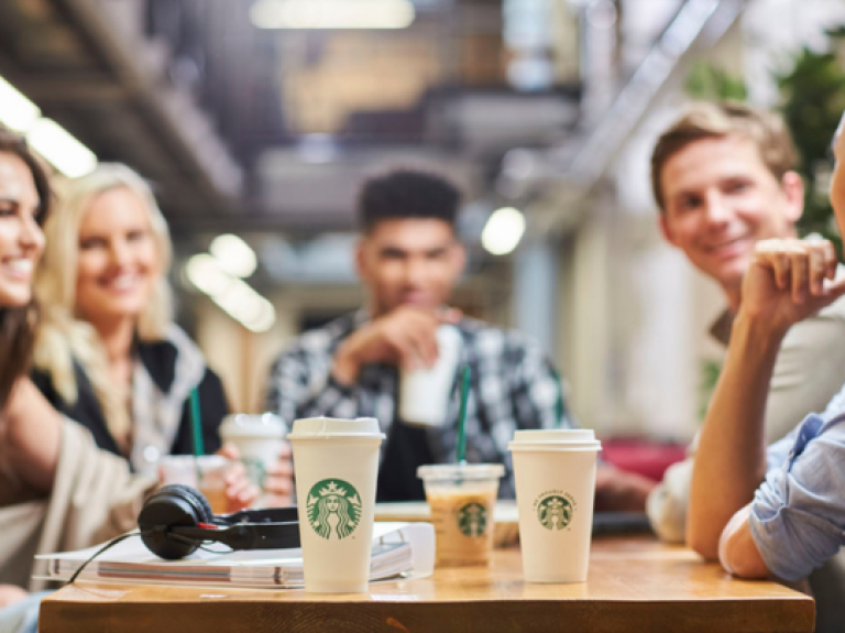 Group of friends gathered around a table with Starbucks coffee cups and headphones in a casual indoor setting.