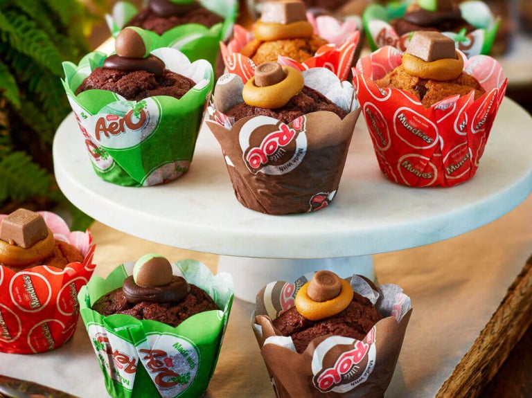 Assorted chocolate and caramel muffins topped with candies, displayed on a white marble stand and wooden tray.