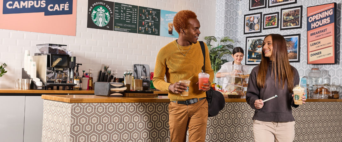 Two people holding Starbucks drinks chat while leaving a patterned campus café with a barista behind the counter.