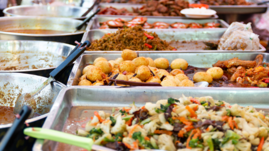 Variety of cooked dishes including vegetables, fried snacks, and rice served in metal trays at a market food stall.