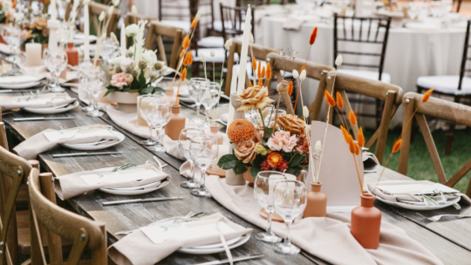 Long wooden table set for event with beige napkins, glassware, floral centerpieces, dried orange accents, and candles