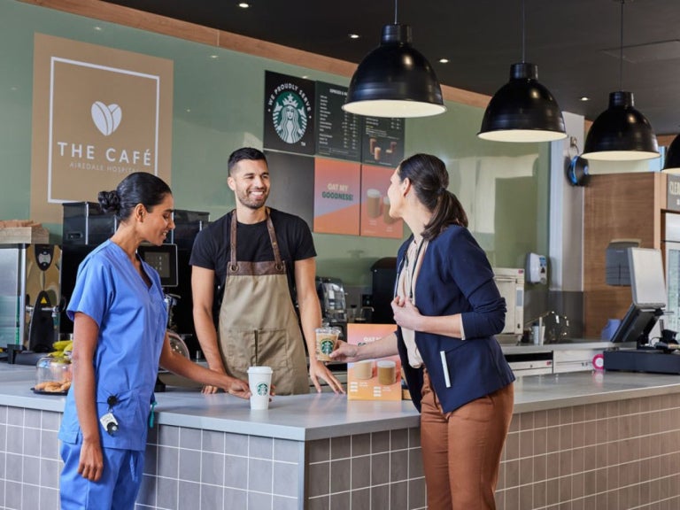 Two women and a barista at a café counter, with one woman wearing scrubs and the other taking a Starbucks drink.
