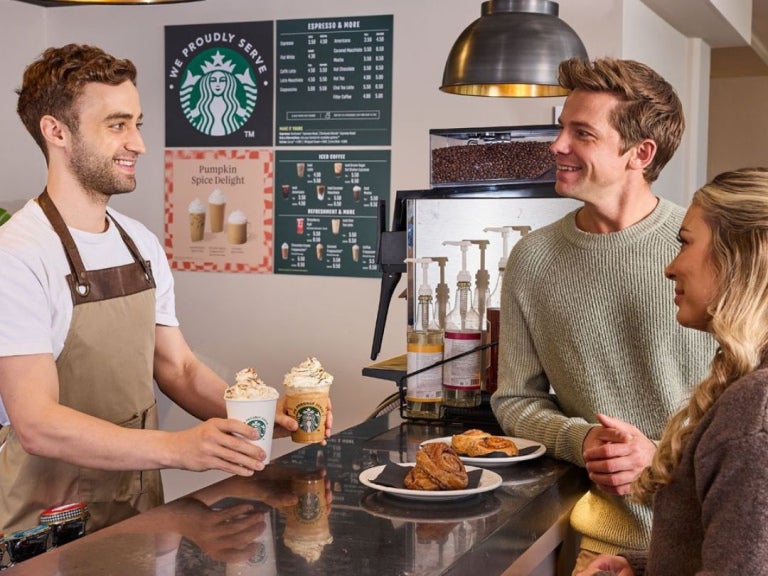 Barista serves two whipped cream coffee drinks to customers at a Starbucks counter with pastries and menu boards visible.