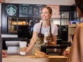 Barista in apron handing two Starbucks coffee cups to customers at a coffee shop counter with menu boards in the background.