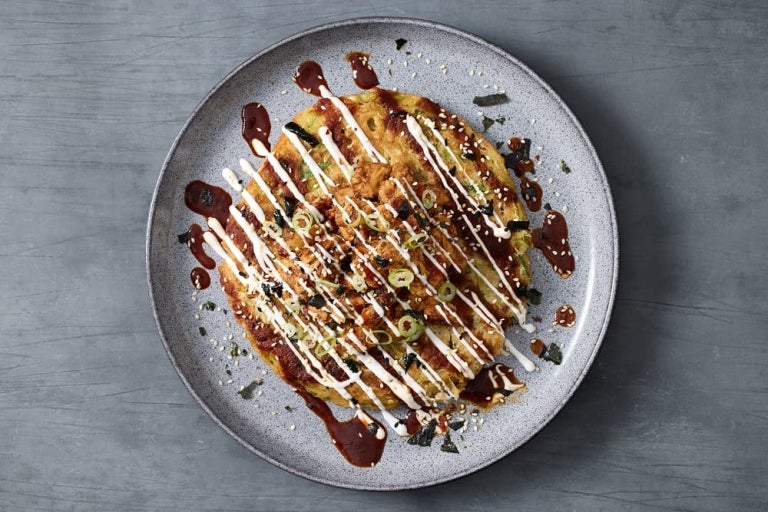 A beautifully arranged okonomiyaki topped with drizzles of sauce, mayonnaise, green onions on a gray plate.