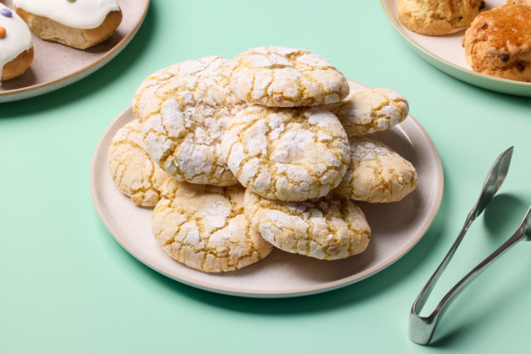 Plate of powdered sugar-dusted lemon crinkle cookies on a light green table with pastry tongs nearby.