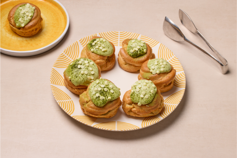 Plate of six cream puffs topped with green matcha glaze and white chocolate chips, with tongs and an additional puff