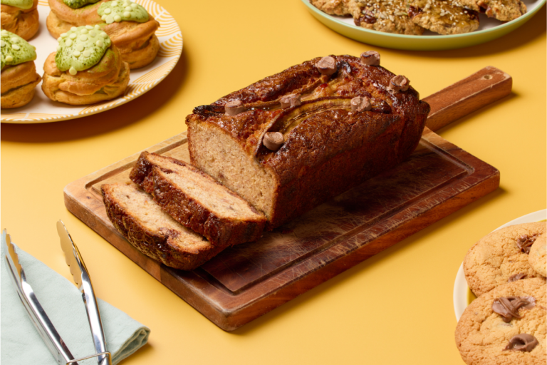 Sliced banana bread with chocolate chips on a wooden board, surrounded by cookies, cream puffs with green icing, and oatmeal 