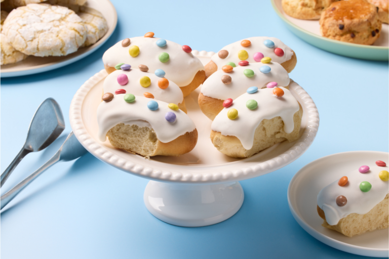 Plate of sweet bread rolls with white icing and colorful candy toppings on a white cake stand against a blue background.