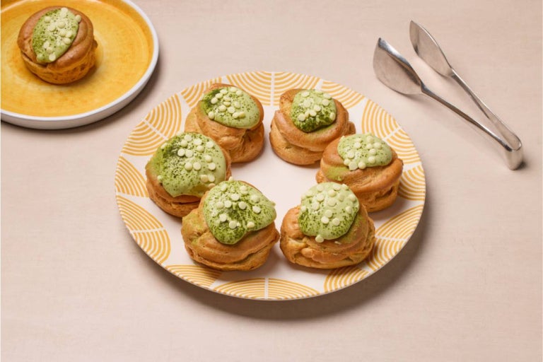 Plate of six cream puffs topped with green matcha glaze and white chocolate chips, with tongs and an additional puff