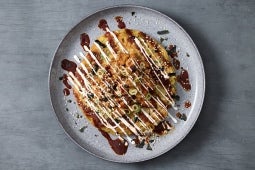 A beautifully arranged okonomiyaki topped with drizzles of sauce, mayonnaise, green onions on a gray plate.