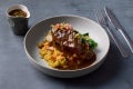 A plated dish featuring tender meat topped with sauce, garnished with vegetables and herbs