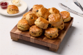 A wooden board stacked with freshly baked golden-brown scones with raisins, with a plate of scone, jam, and cream in the back