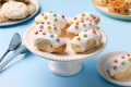 Plate of sweet bread rolls with white icing and colorful candy toppings on a white cake stand against a blue background.