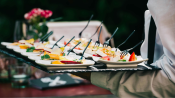 Waiter carrying tray with multiple plates of gourmet appetizers, each with a small fork, against a blurred background.