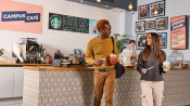 Two people holding Starbucks drinks chat while leaving a patterned campus café with a barista behind the counter.