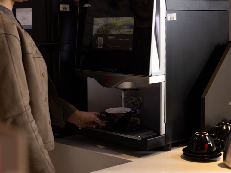 A person pours coffee from a Nescafé machine into a black cup, with coffee capsules and cups arranged nearby.