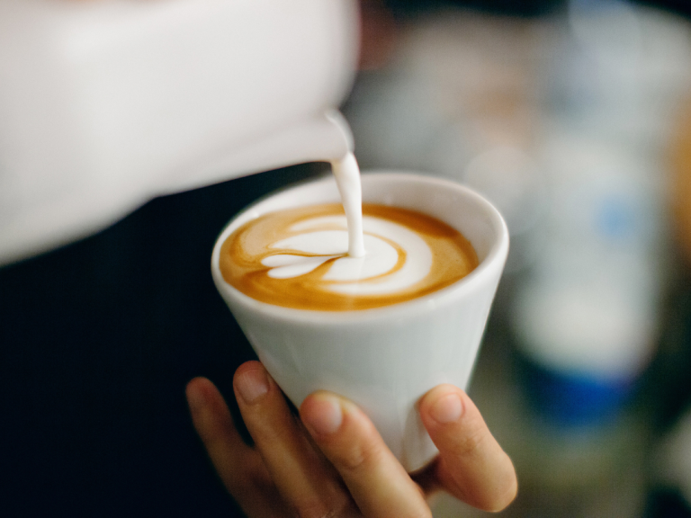 Close-up of hand holding white cup while pouring steamed milk to create latte art on coffee.