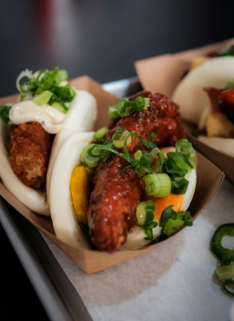 Close-up of bao buns filled with fried chicken, pickles, scallions, and mayo served in brown paper trays.
