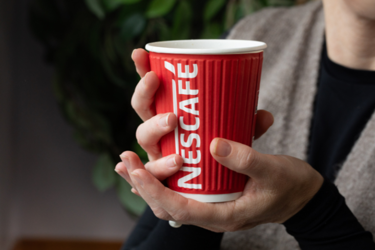 Person holding a red Nescafé paper cup with both hands against a blurred indoor background.